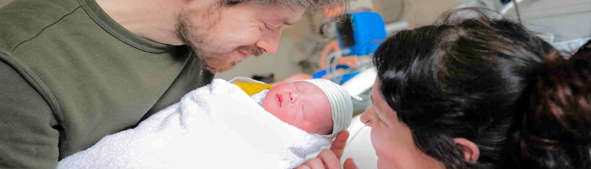 A smiling man and woman look at a newborn baby with medical equipment in the background A smiling man and woman look at a newborn baby with medical equipment in the background